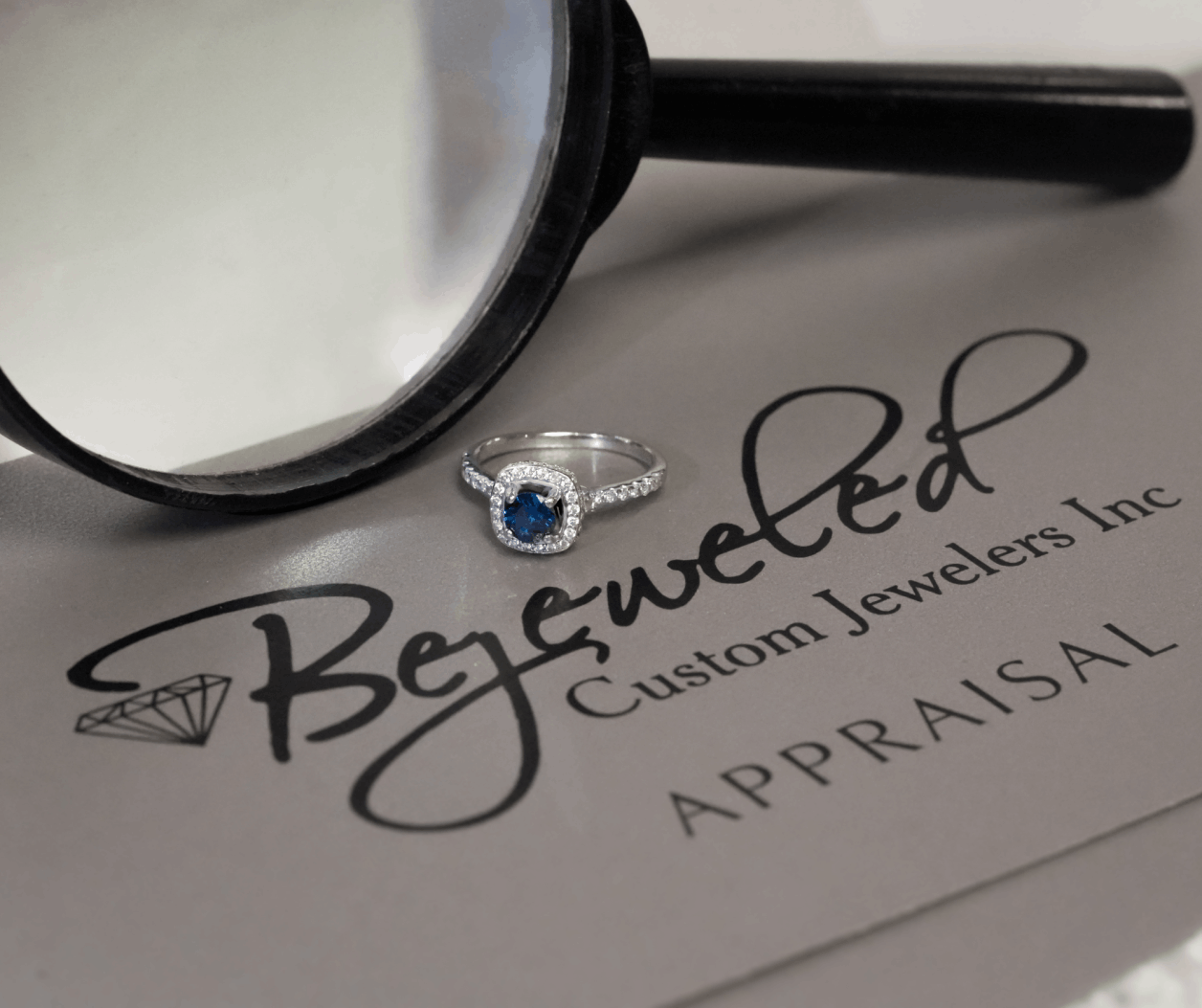 A silver or white gold ring with a square-cut blue sapphire and diamond halo resting on a gray appraisal folder from Bejeweled Custom Jewelers Inc, next to a magnifying glass
