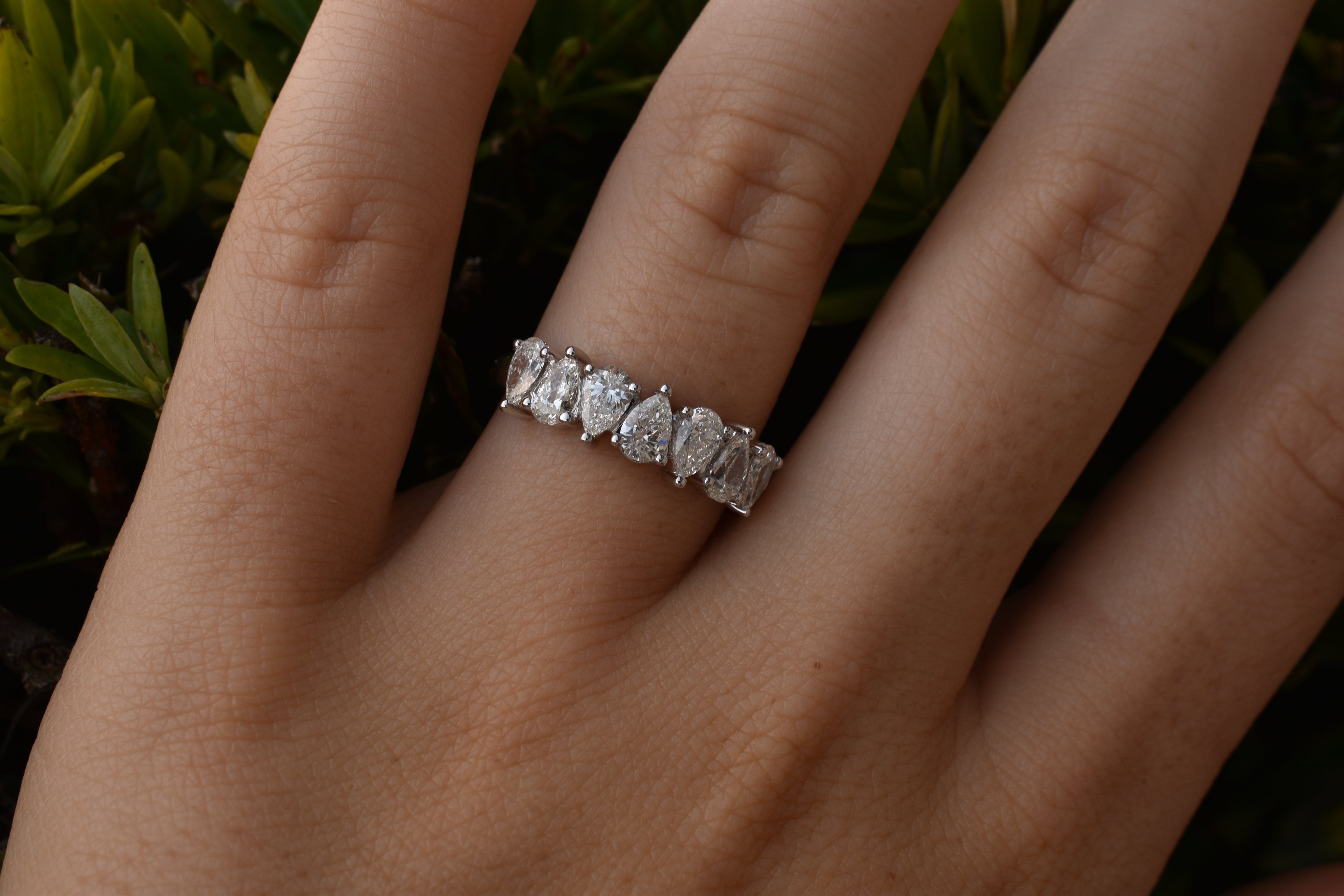 Silver or white gold ring featuring a horizontal row of seven pear-shaped diamonds, worn on a person's finger against a green foliage background