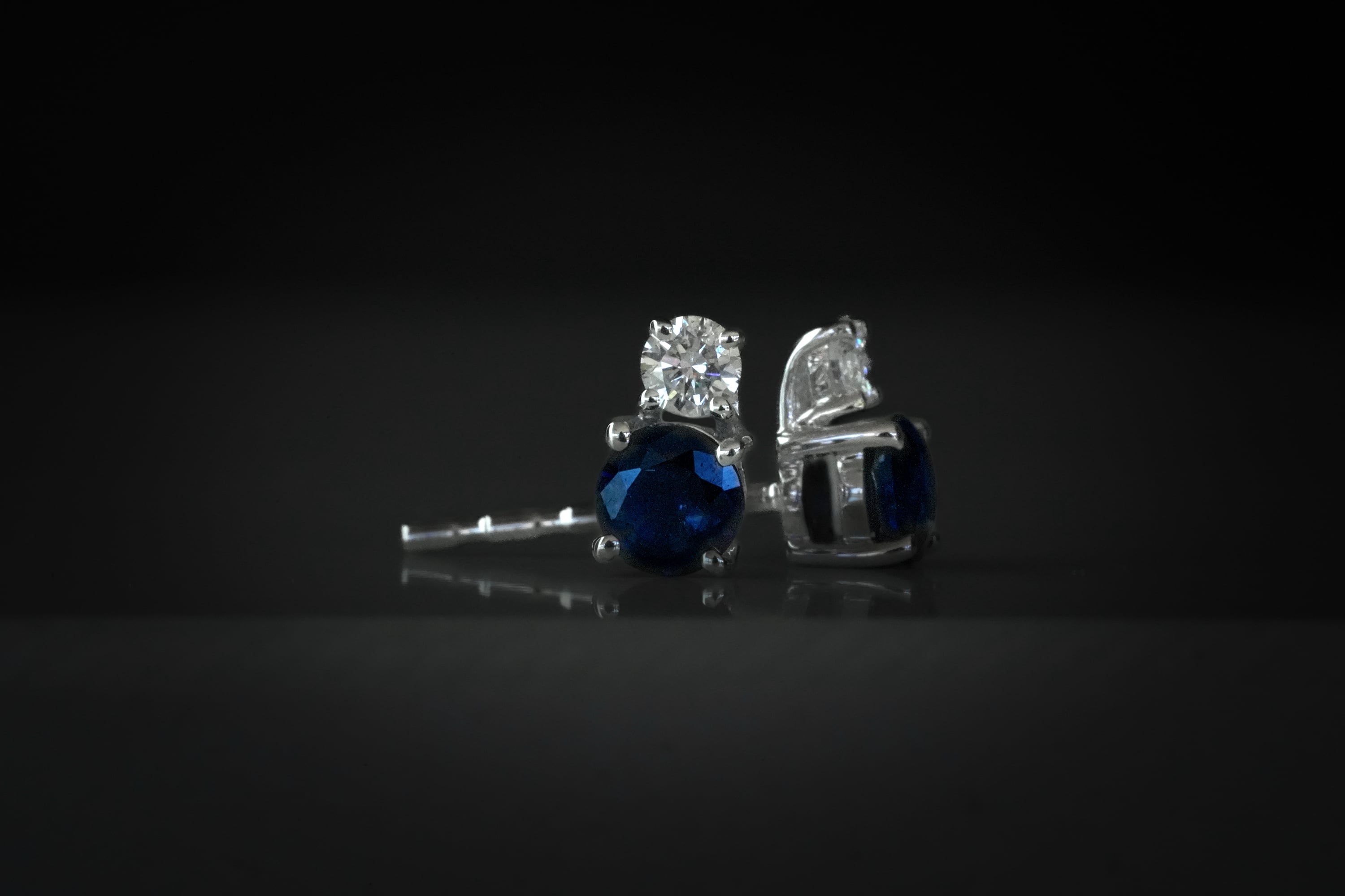 Silver stud earrings featuring a round dark blue sapphire topped with a smaller brilliant round diamond, shown against a dark, reflective background