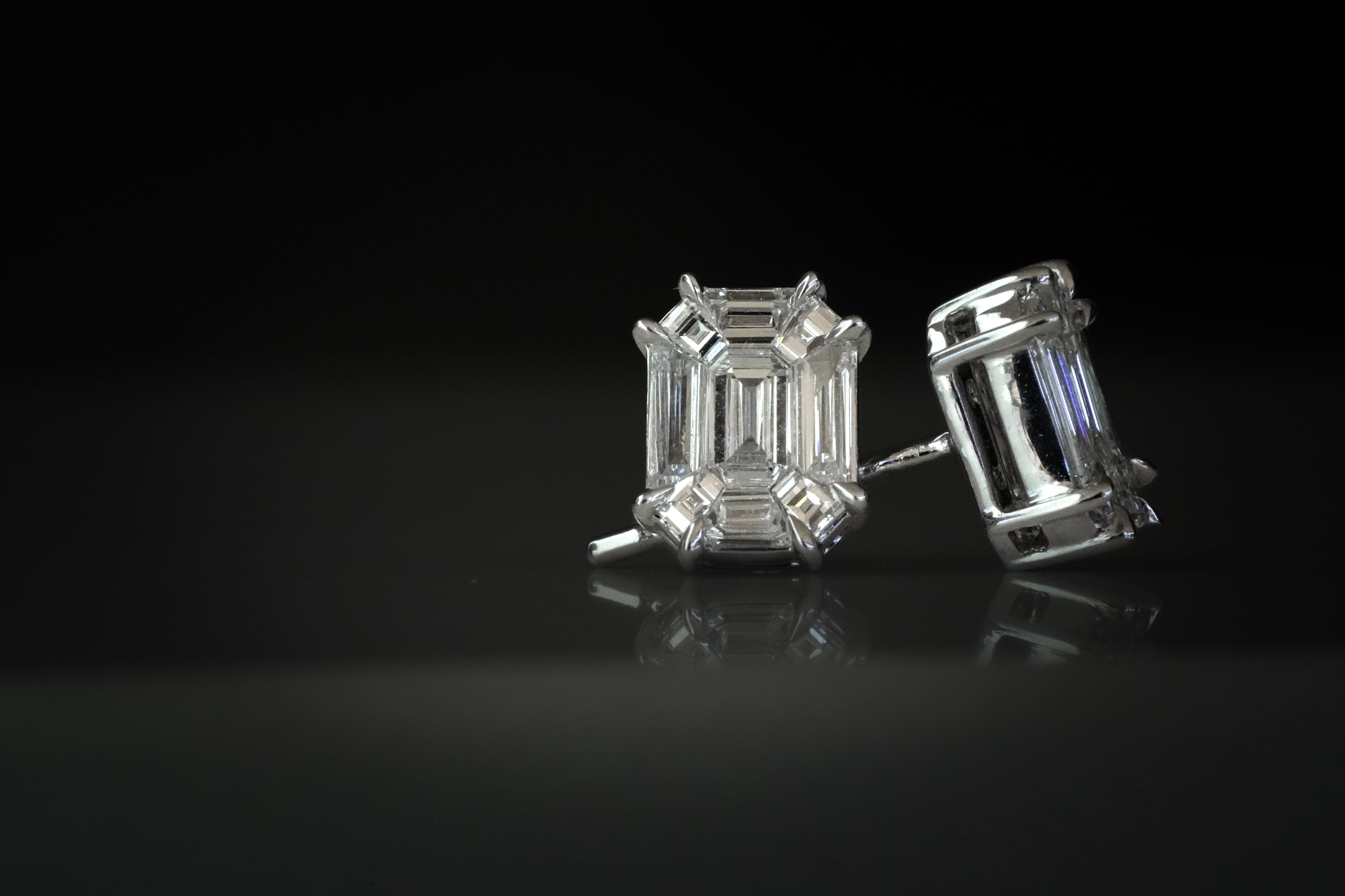 Silver or white gold stud earrings featuring large emerald-cut diamonds in a four-prong setting, displayed on a dark reflective surface