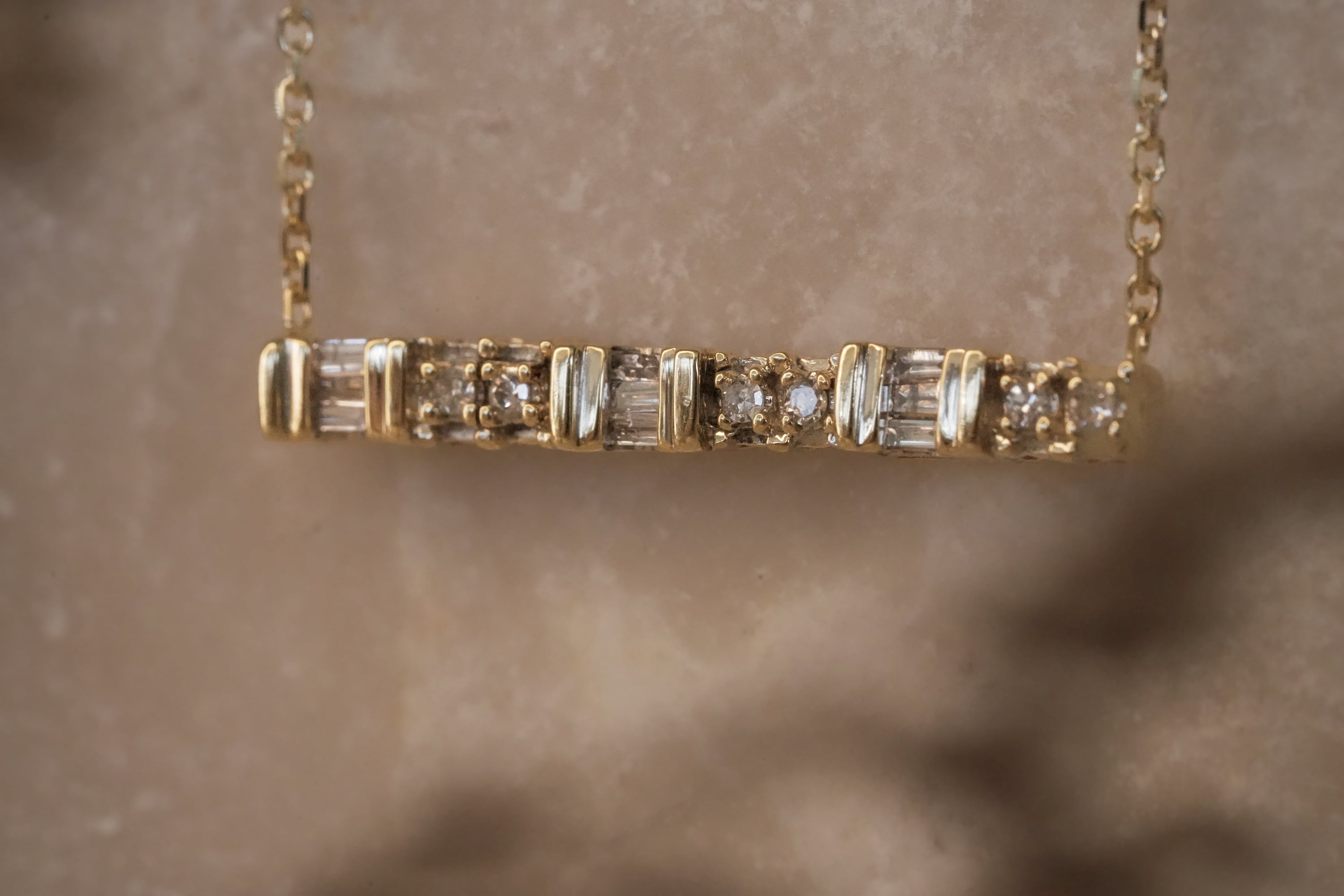 Gold bar necklace featuring alternating baguette and round diamonds in a channel setting, suspended from a fine gold chain against a stone background