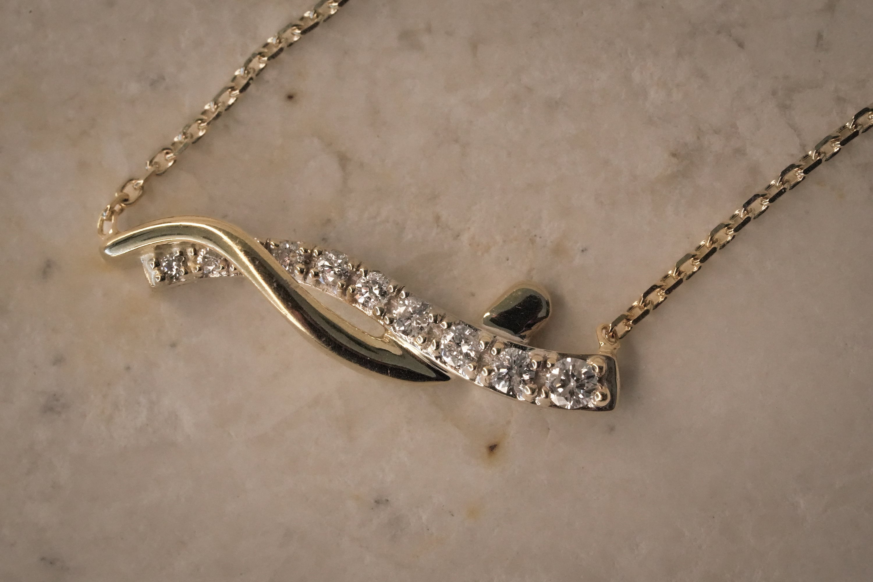 Gold wave necklace featuring a curved row of round diamonds intertwined with a polished gold ribbon, displayed on a light-colored stone background