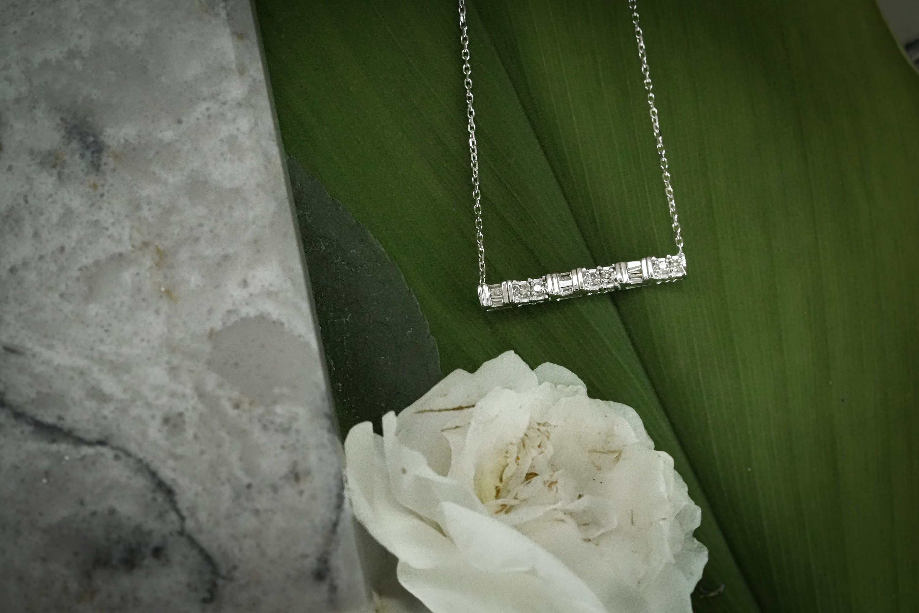 A silver or white gold bar necklace featuring a row of baguette and round-cut diamonds, laid across a large green leaf next to a white flower and a marble surface