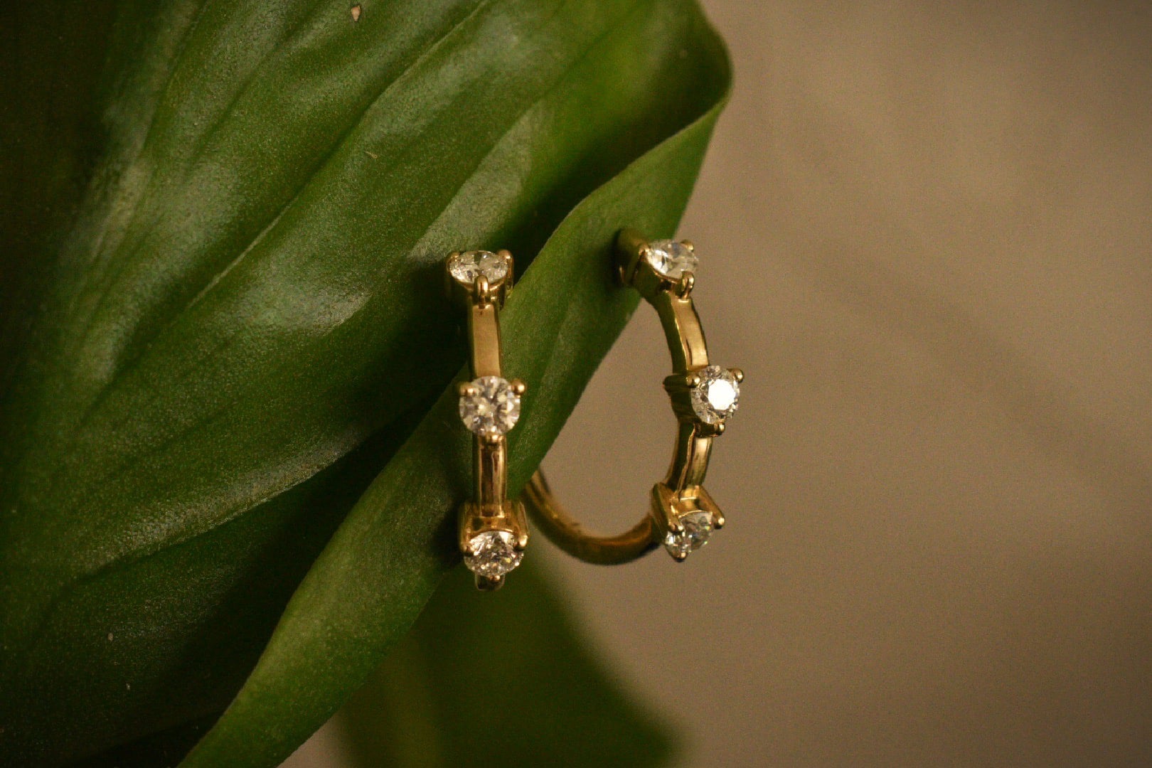 Gold hoop earrings featuring three round brilliant-cut diamonds in vertical prong settings, hooked through a vibrant green leaf on a neutral background